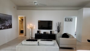 A modern living room with a white sofa, a smaller armchair, a flat-screen TV, a ceiling fan, and a decorative light fixture.
