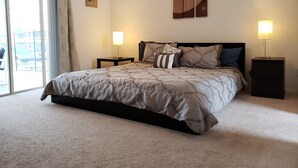 A bedroom with a large bed, two bedside tables, a nightstand, a wall-mounted lamp, and a sliding glass door leading to an outdoor area.