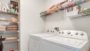 Un cuarto de lavandería con dos lavadoras de carga frontal apiladas, repisas con toallas dobladas y una cesta de productos de limpieza.