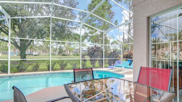 Zona de piscina con cerramiento de vidrio, con una mesa y sillas, rodeada de una estructura de metal blanco y vistas a un jardín ajardinado.