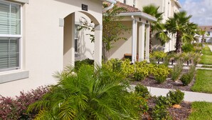A two-story house with a beige exterior, a number '1574' on the side, and a well-maintained garden with various plants and a palm tree. - Champions Gate 1570 (Davenport)