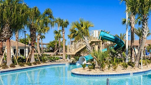 A swimming pool with a water slide, palm trees, and a building in the background. - Champions Gate 1570 (Davenport)