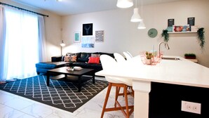 A modern living room with a black sofa, a coffee table, and a kitchen area with a white countertop and bar stools. - Champions Gate 1570 (Davenport)
