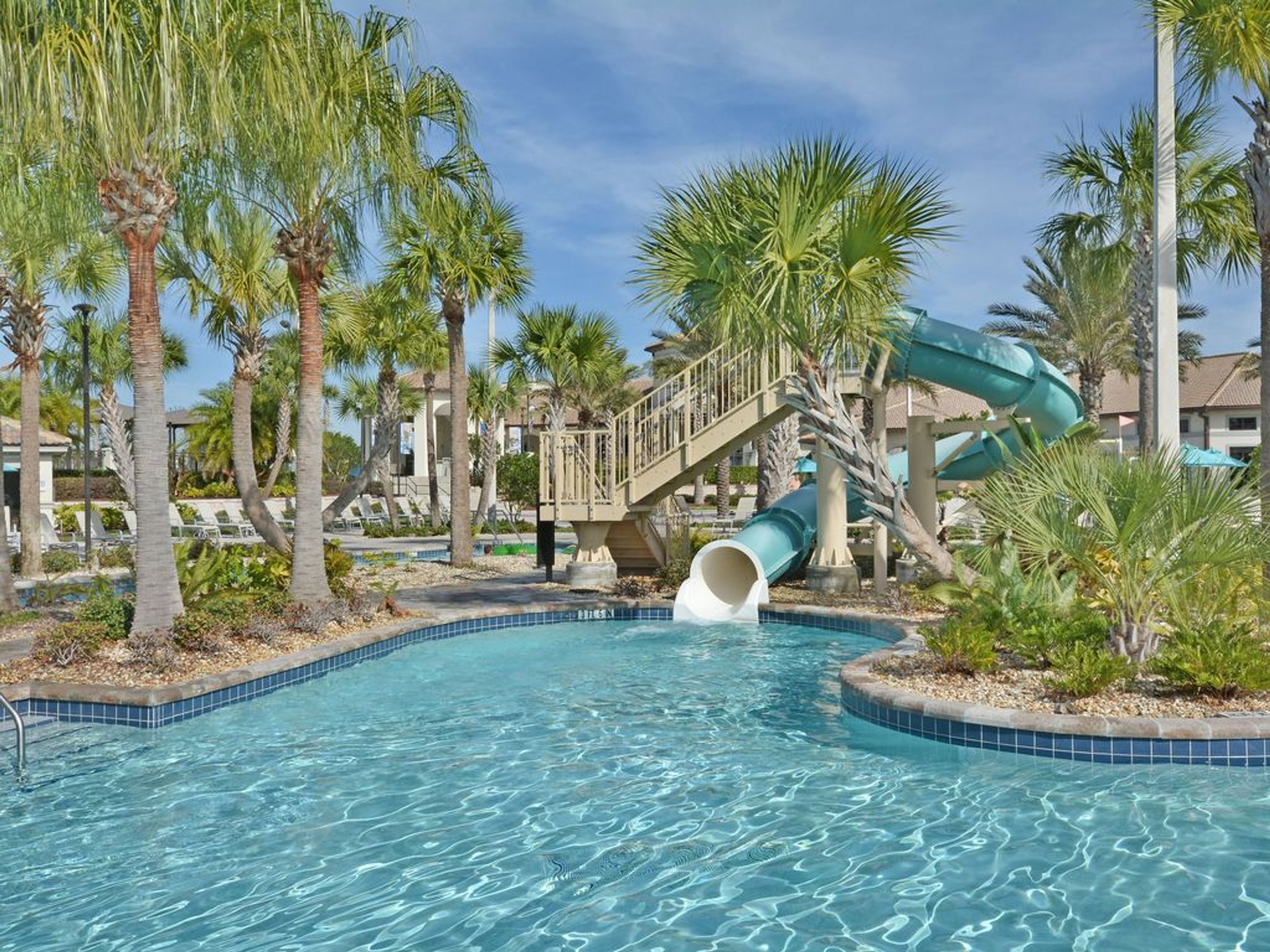 A swimming pool with a water slide, palm trees, and a bridge.