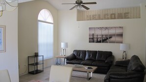 A living room with a brown leather sofa, a coffee table, a dining table with plates and a wine glass, a lamp, and a 'WELCOME' sign on the wall. - Indian Wells 3120 (Kissimmee)