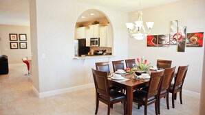Comedor con una mesa dispuesta para seis personas, sillas alrededor y una cocina visible a través de un arco.