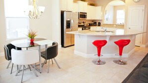 Una cocina moderna con gabinetes blancos, electrodomésticos de acero inoxidable, una isla central con taburetes y una zona de comedor con mesa y sillas.