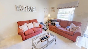 A living room with two red sofas, a glass coffee table, and a 'HOME' sign on the wall.