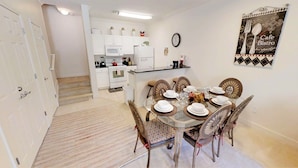 A dining area with a glass table set for six, surrounded by six chairs with patterned cushions. A kitchen with white cabinets and appliances is visible in the background.