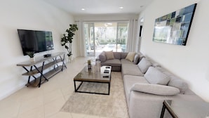 A modern living room with a large flat-screen TV, a sofa, a coffee table, and a dining area with a plant. - Encore Resort at Reunion 7511 (Kissimmee)