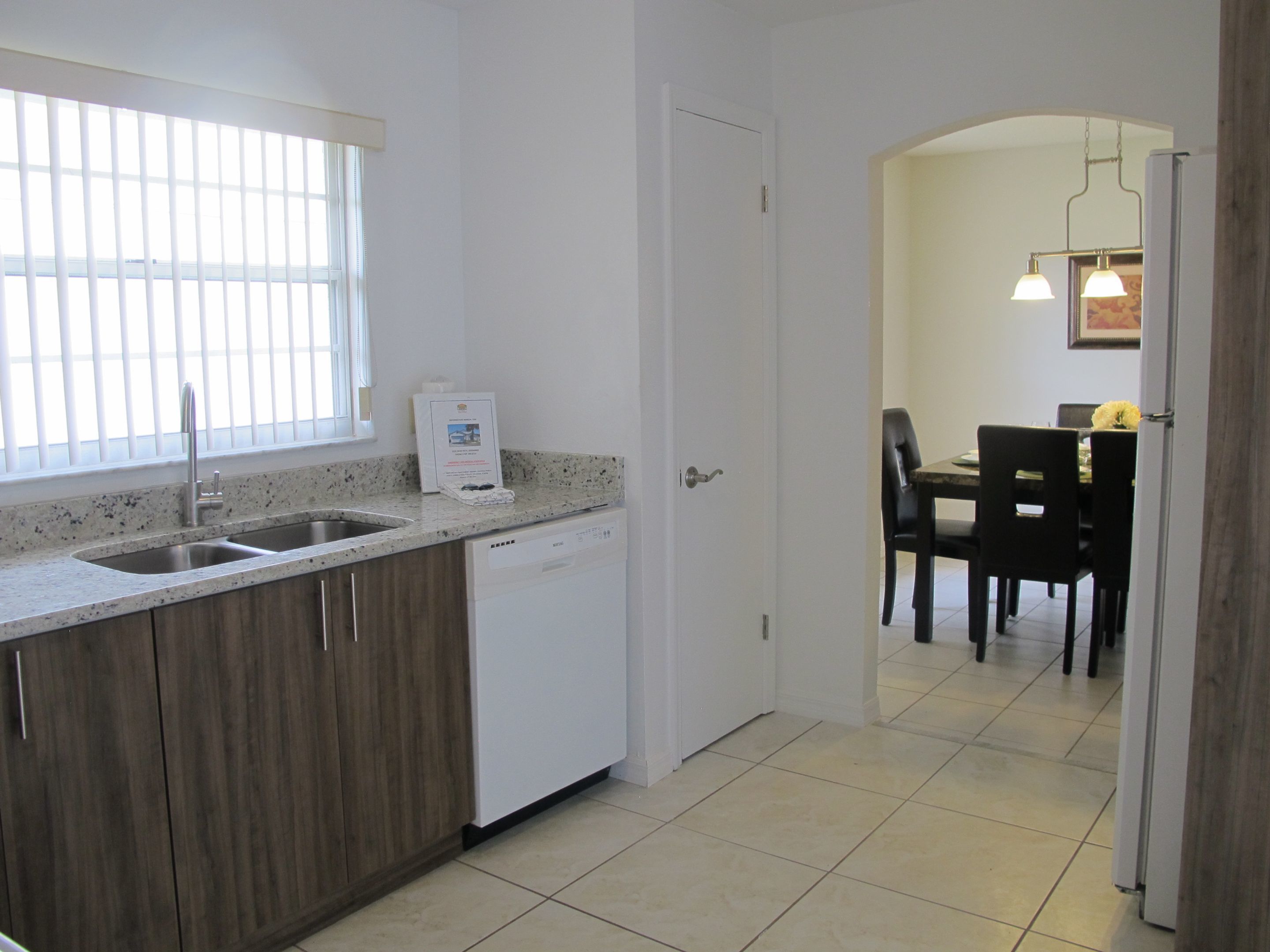 A kitchen with a dishwasher, a window with blinds, and a dining area with a table and chairs.