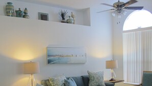 Sala de estar con dos sofás, una mesa de centro y un ventilador de techo. Hay un cuadro en la pared y una ventana con persianas.