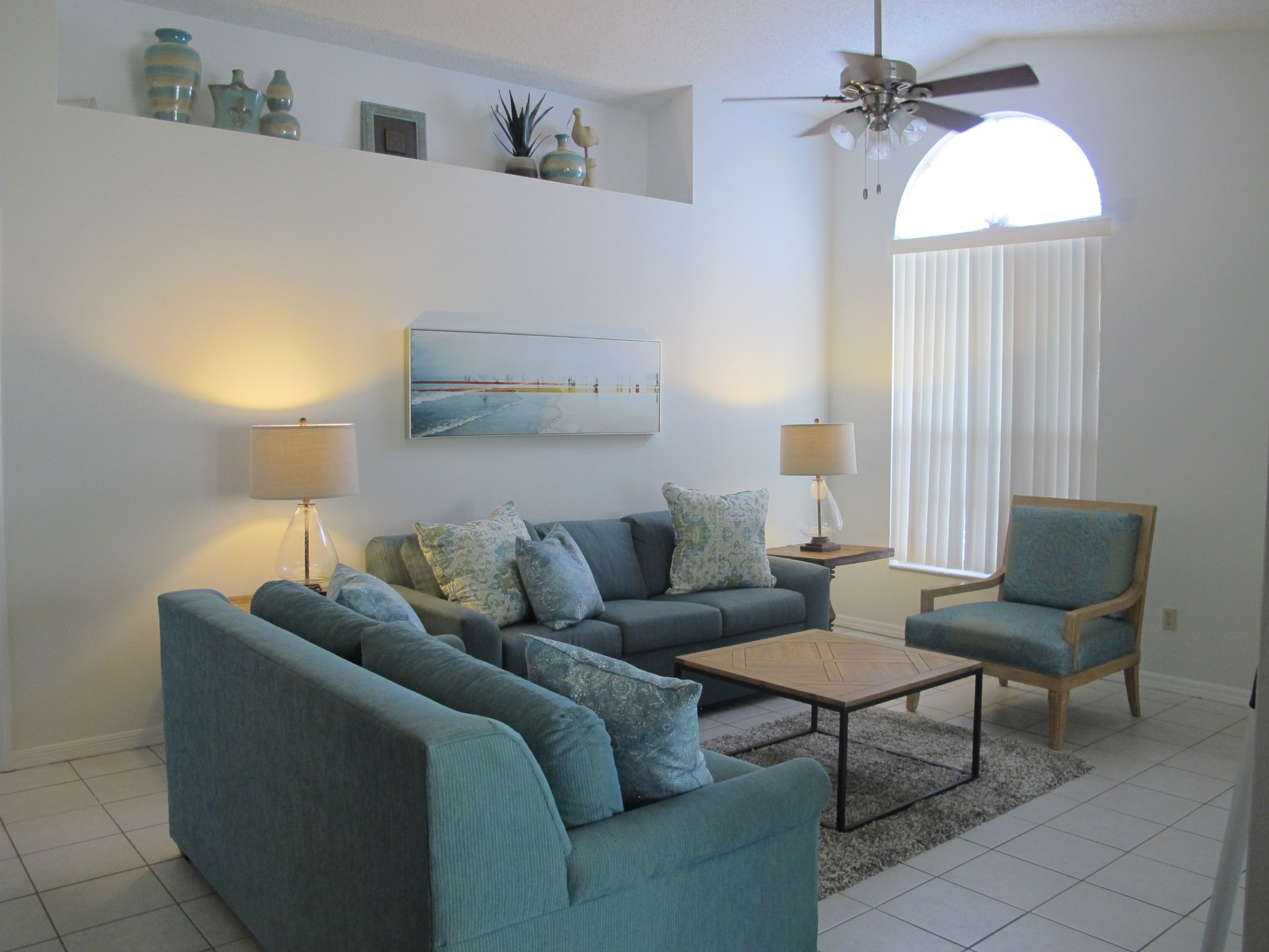 A living room with a sofa set, two side tables with lamps, a coffee table, a chair, a ceiling fan, and a large window with blinds.