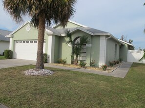 A single-story house with a green exterior, a white garage door, and a small front yard with a palm tree and various plants. - Indian Wells 3133 (Kissimmee)