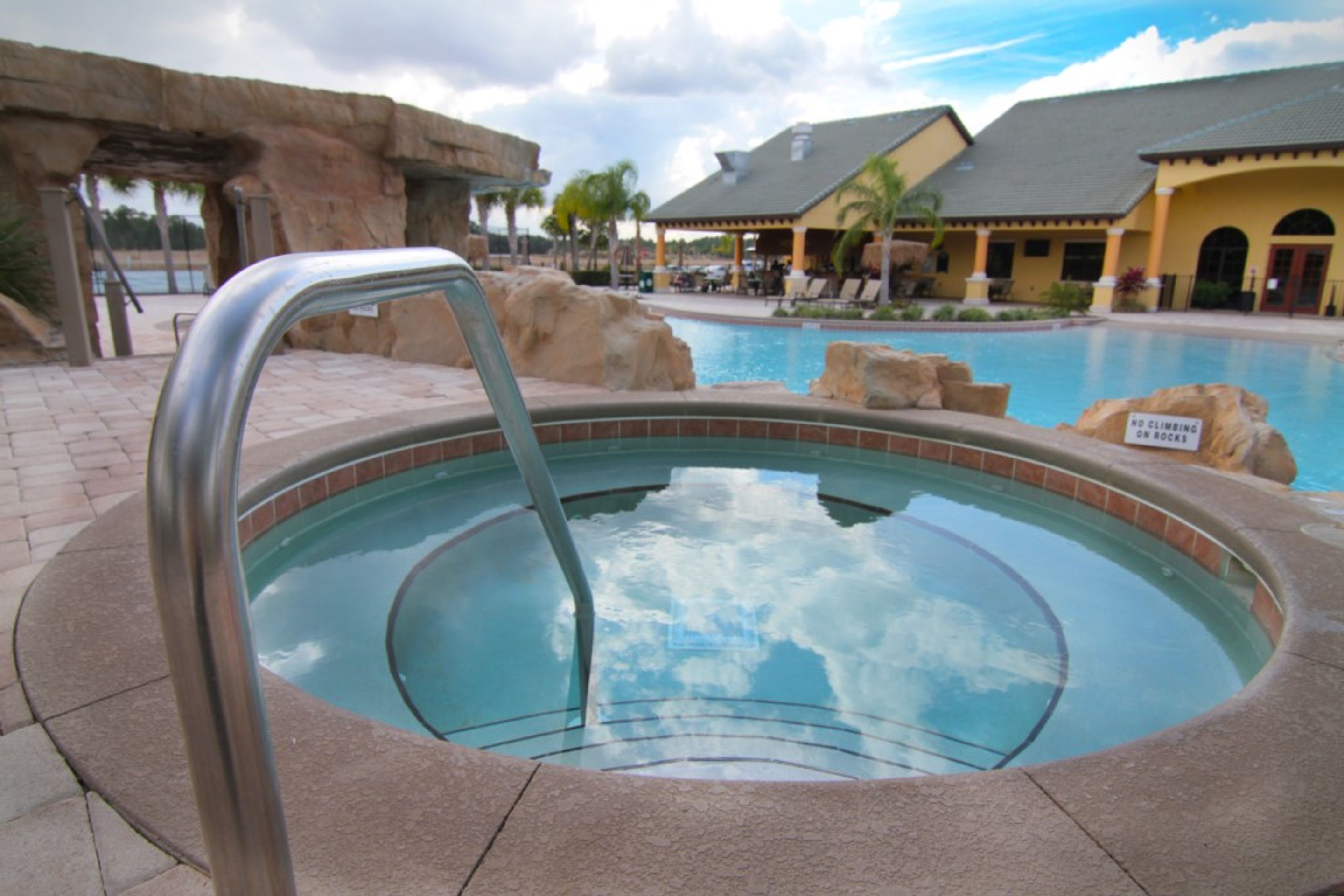 A hot tub with a metal railing, surrounded by a pool area with lounge chairs and a building in the background.