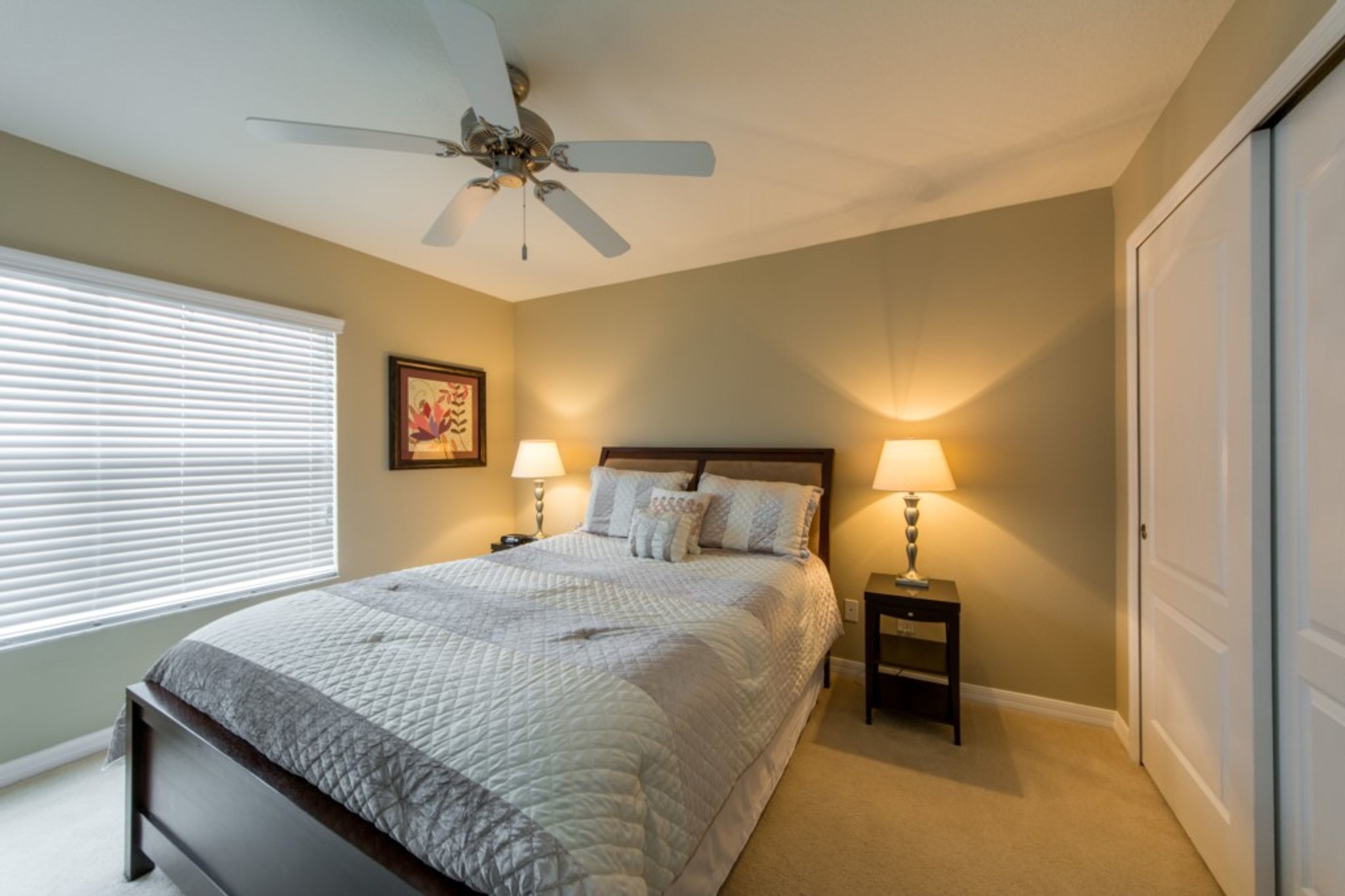 A bedroom with a bed, two bedside tables with lamps, a ceiling fan, a window with blinds, and a door.
