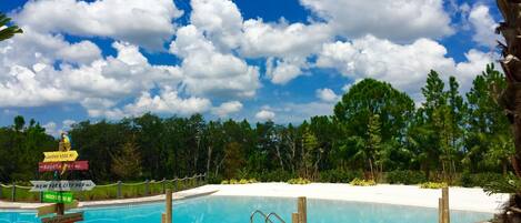 A swimming pool with a tiled deck, surrounded by palm trees and a clear blue sky with clouds.