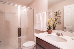 Une salle de bain avec une douche en verre, des toilettes blanches, un lavabo avec un miroir et une plante en pot aux fleurs jaunes.