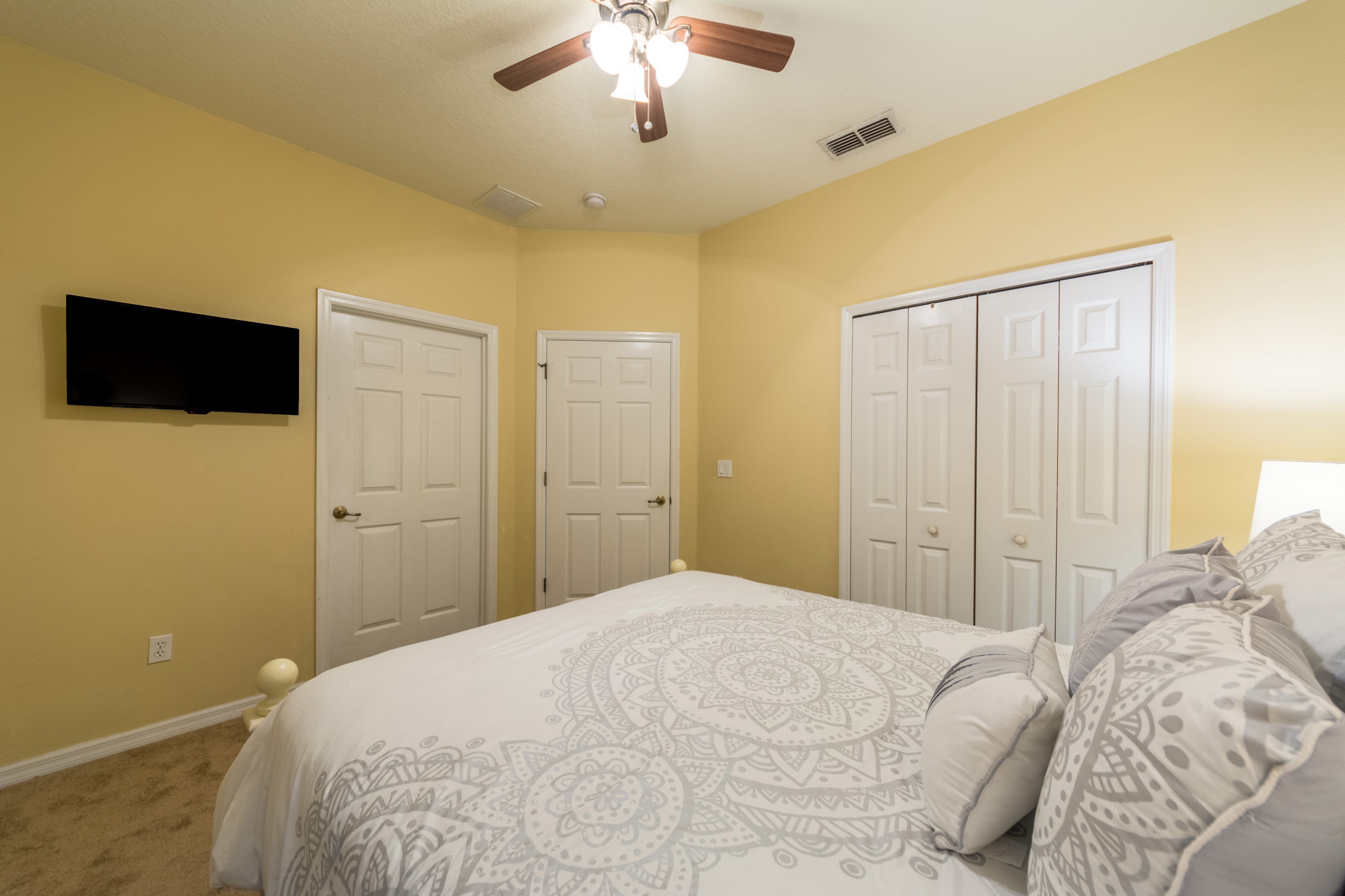 A bedroom with a bed, two doors, a ceiling fan, and a mounted TV.