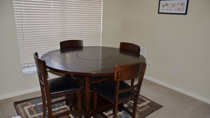 Een eetgedeelte met een ronde tafel, vier stoelen, een kamerbreed tapijt en een raam met jaloezieën.
