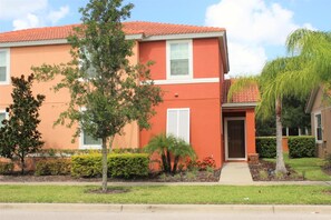 A two-story building with a red exterior, white trim, and a brown door. There are trees and shrubs in front of the building, and a sidewalk leading to the entrance. - Bella Vida 540 (Kissimmee)