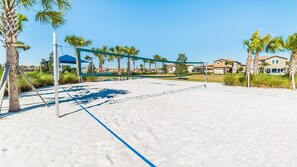 A beach volleyball court with a net, palm trees, and houses in the background.