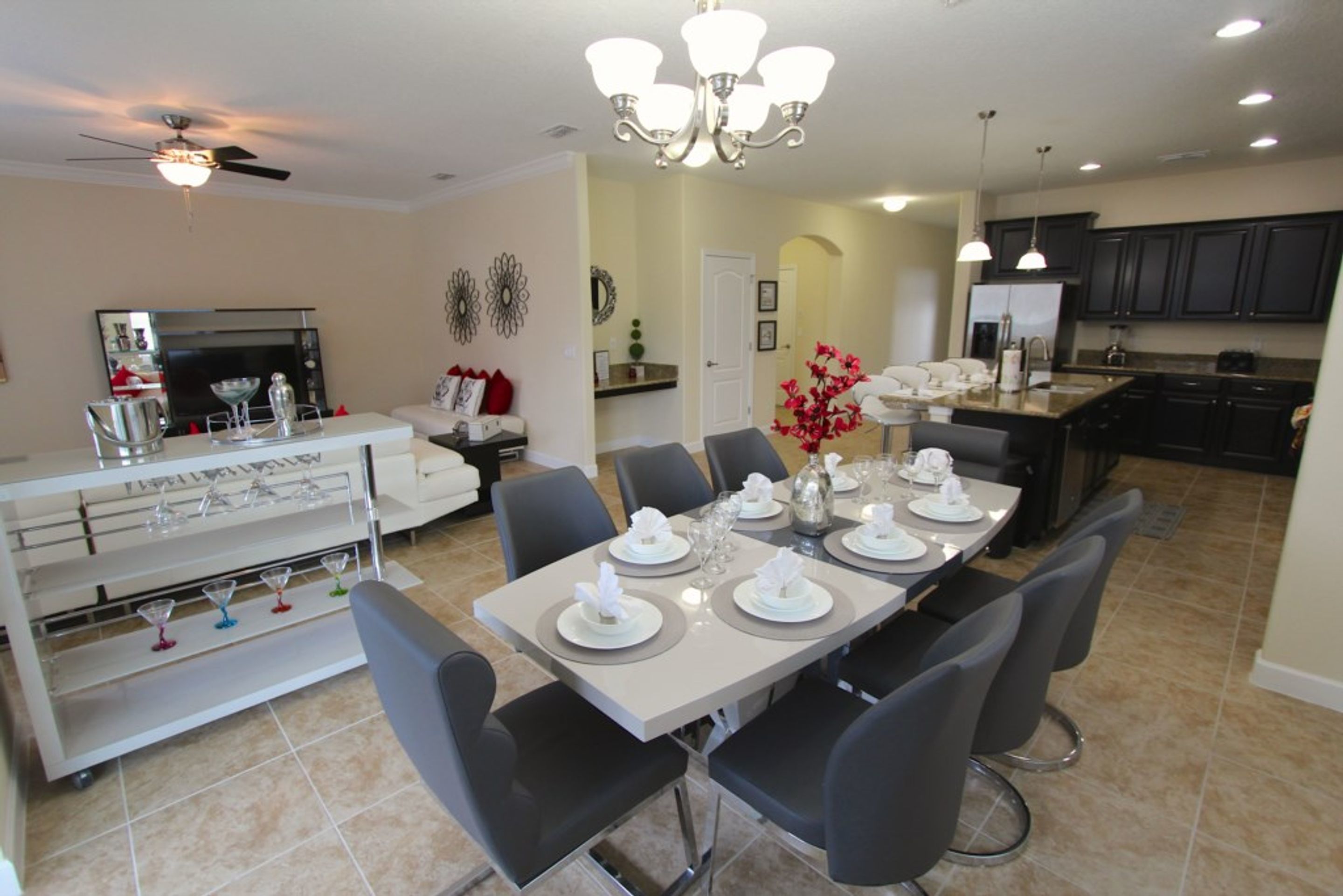 A dining area with a long table set for six, surrounded by chairs. A chandelier hangs above, and a ceiling fan is visible. The room has a modern design with a kitchen in the background.