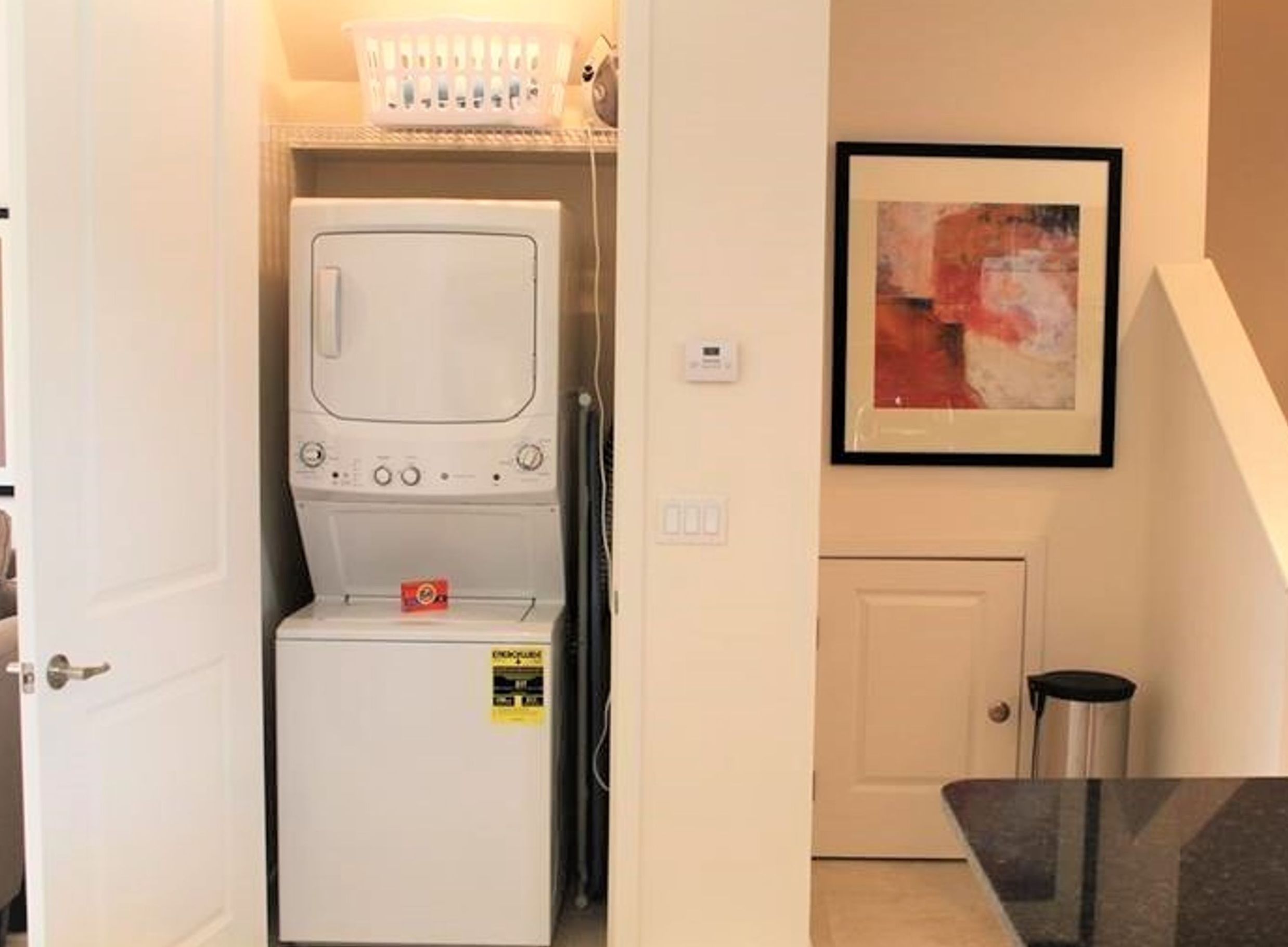 A laundry room with a stacked washer and dryer, a trash can, and a framed abstract painting on the wall.