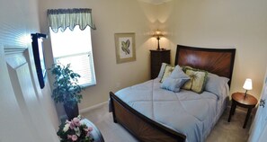 A bedroom with a bed, a nightstand, a lamp, a window with blinds, a plant, and a framed picture on the wall.