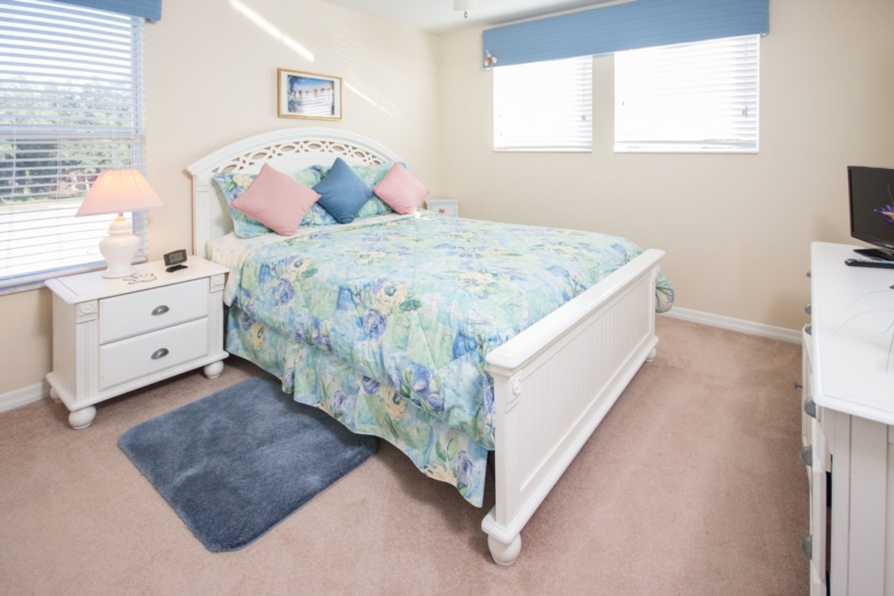 A bedroom with a bed, a nightstand, a lamp, a desk, and a computer.