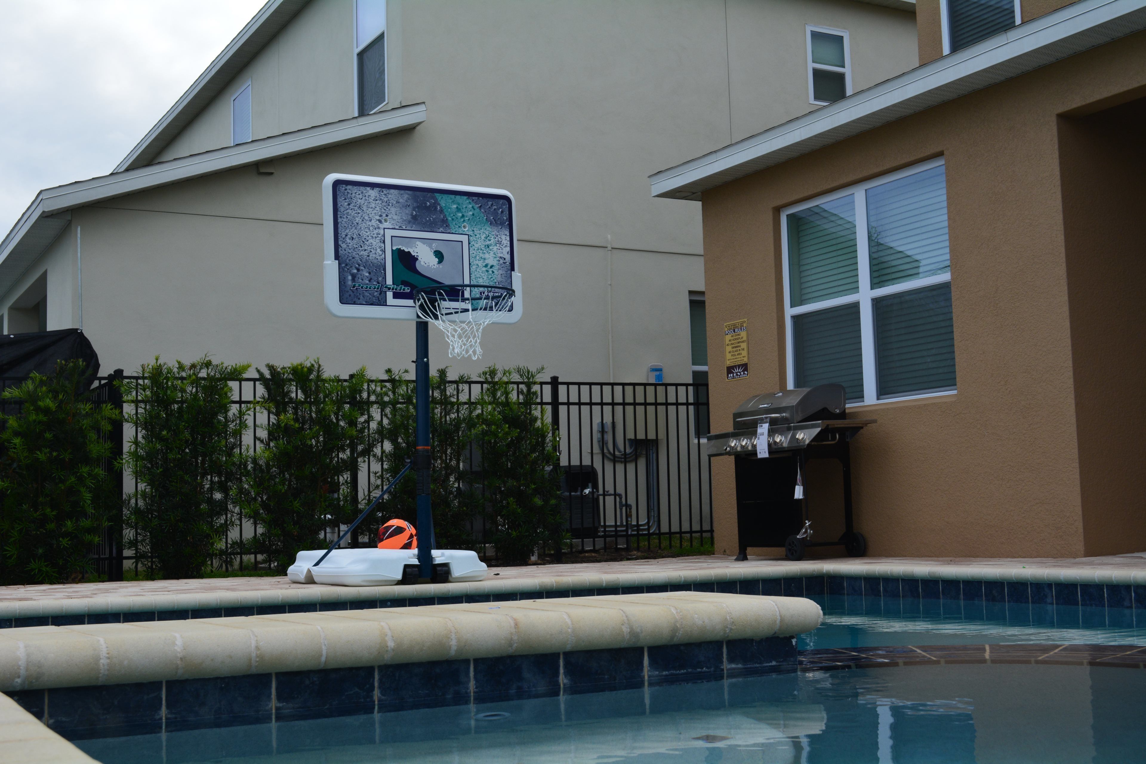 Piscina con aro de baloncesto, rodeada por una cerca y follaje.