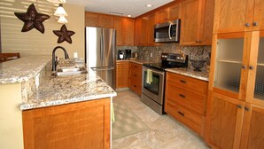 A kitchen with wooden cabinets, stainless steel appliances, granite countertops, and a checkered rug. - Anna Maria Island 1303 - 2 (Bradenton Beach)
