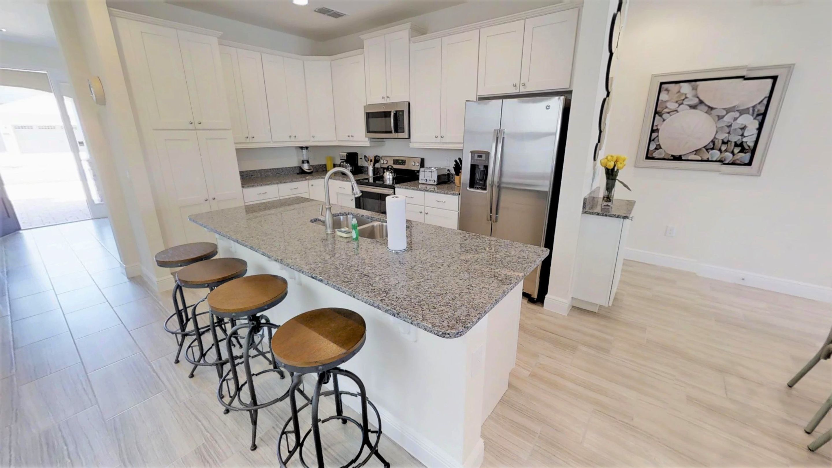 A modern kitchen with white cabinets, a granite countertop, a stainless steel refrigerator, and a microwave. There are four stools at the island.