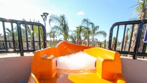A water slide with a yellow structure and a white water flow, surrounded by a metal fence and palm trees. - Solterra 4670 (Davenport)