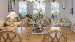 A dining area with a wooden table, chairs, and a floral sofa. There are two windows with curtains, a lamp, and a vase with flowers. - Highlands Reserve Golf & Country Club 533 (Davenport)