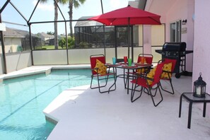 A poolside area with a table and chairs, a red umbrella, and a barbecue grill. - Lake Berkley Resort 4715 B (Kissimmee)