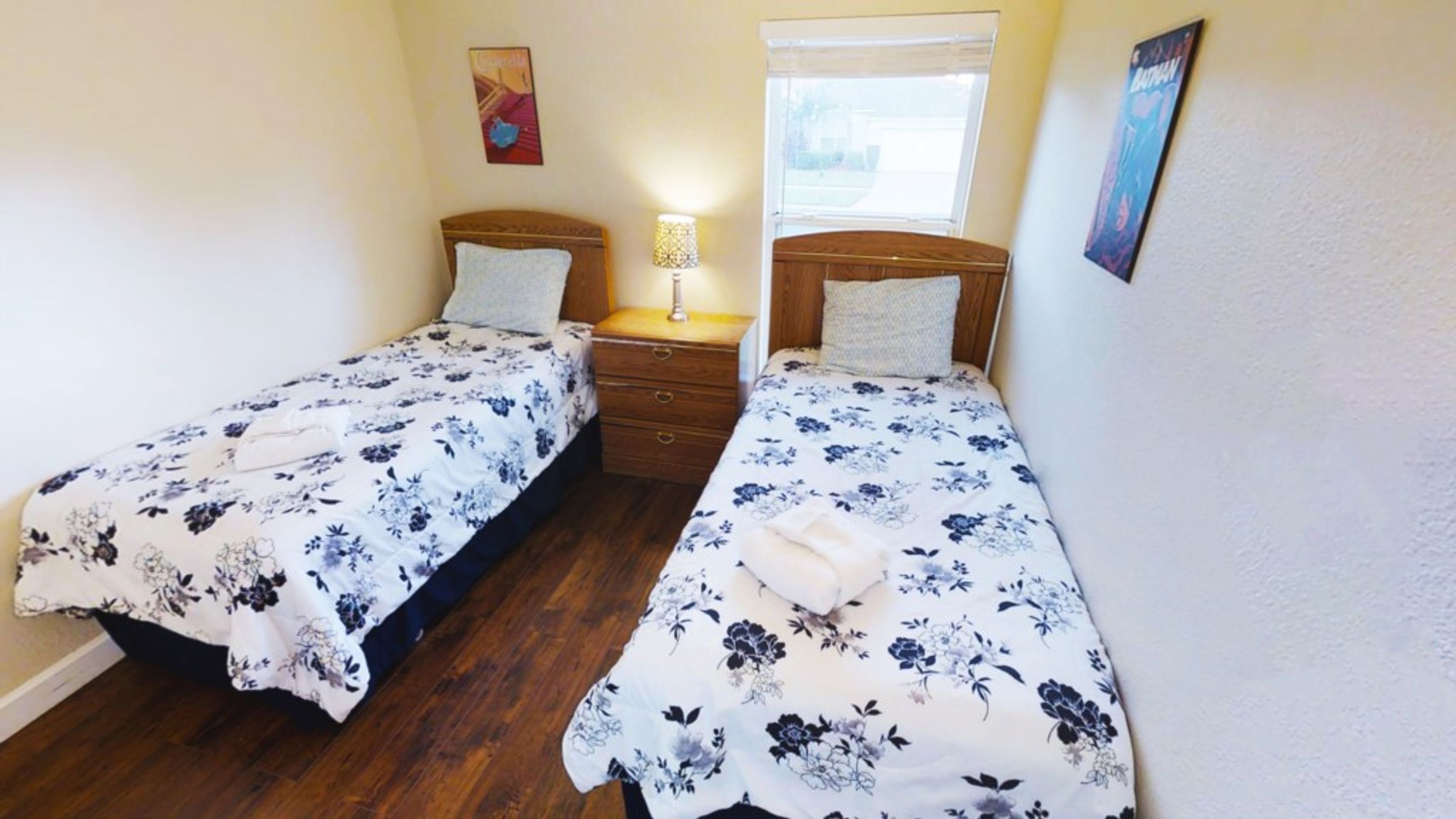 Habitación con dos camas individuales, cada una con colcha estampada y un cojín. Entre las camas hay una mesita de noche de madera con una lámpara. Se ve una ventana con persianas en el fondo.