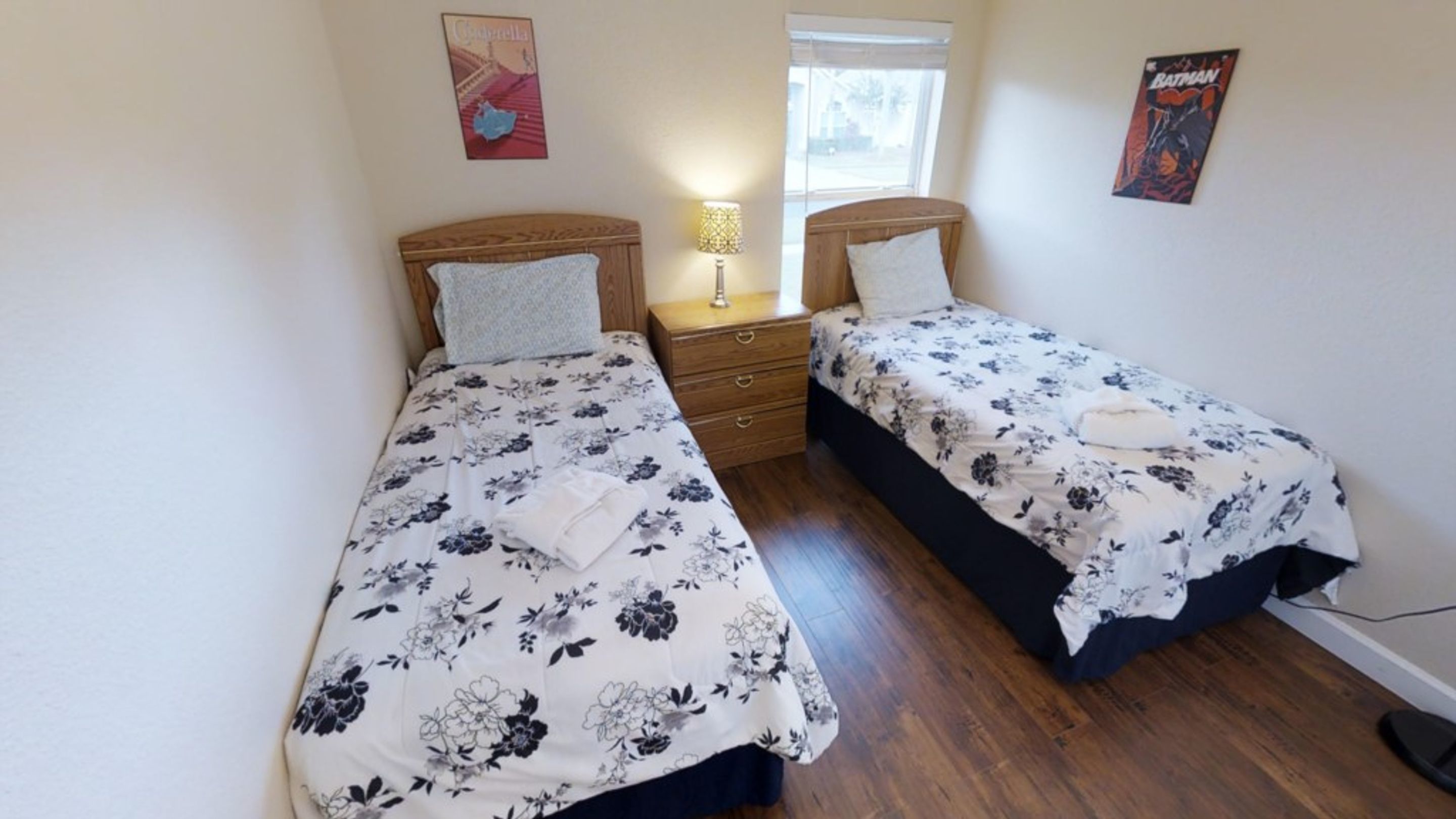 A room with two single beds with floral bedding, a bedside table with a lamp, and a poster on the wall.