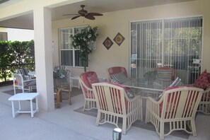 A covered patio area with wicker furniture, a glass table, and a ceiling fan.