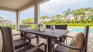 A covered patio area with wicker furniture, a table with drinks, and a view of a swimming pool and palm trees. - Encore Resort at Reunion 281 (Kissimmee)