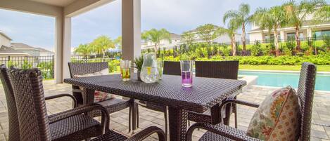 A covered patio area with wicker furniture, a table with drinks, and a view of a swimming pool and palm trees.