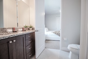 Casa de banho moderna com bancada de granito, armários escuros, sanita branca e quarto visível com cama e porta para outro espaço.