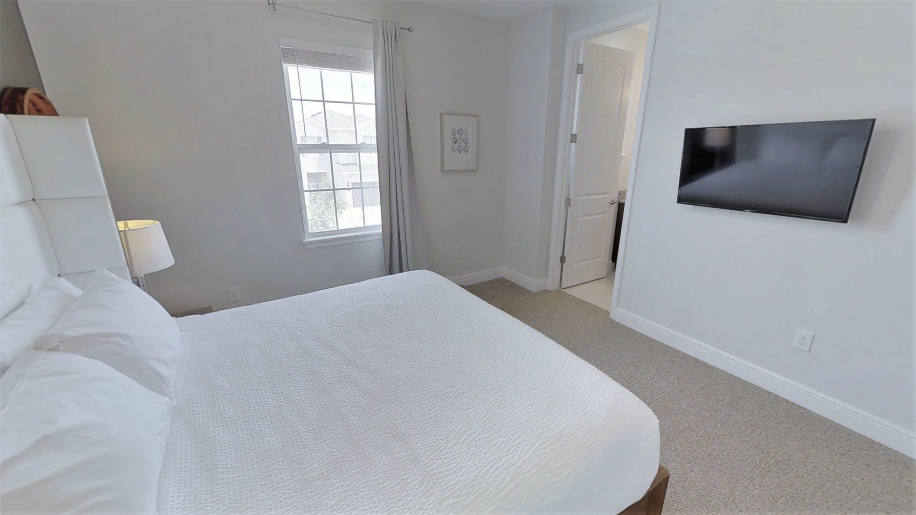 Quarto com cama, televisão fixada na parede, janela com cortinas e porta que dá para outro espaço.