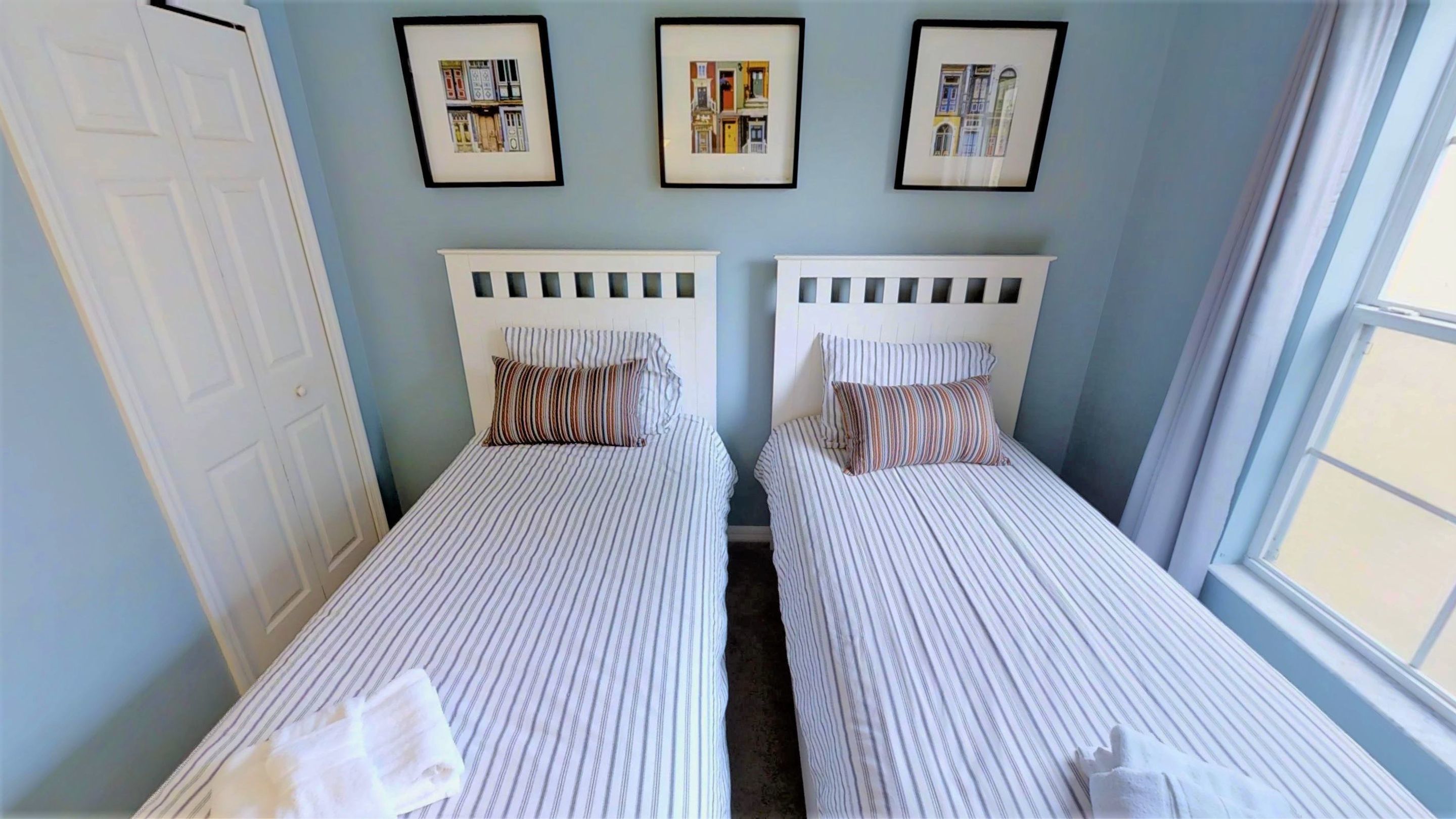 Duas camas de solteiro com roupa de cama listrada, uma porta branca e três quadros na parede.