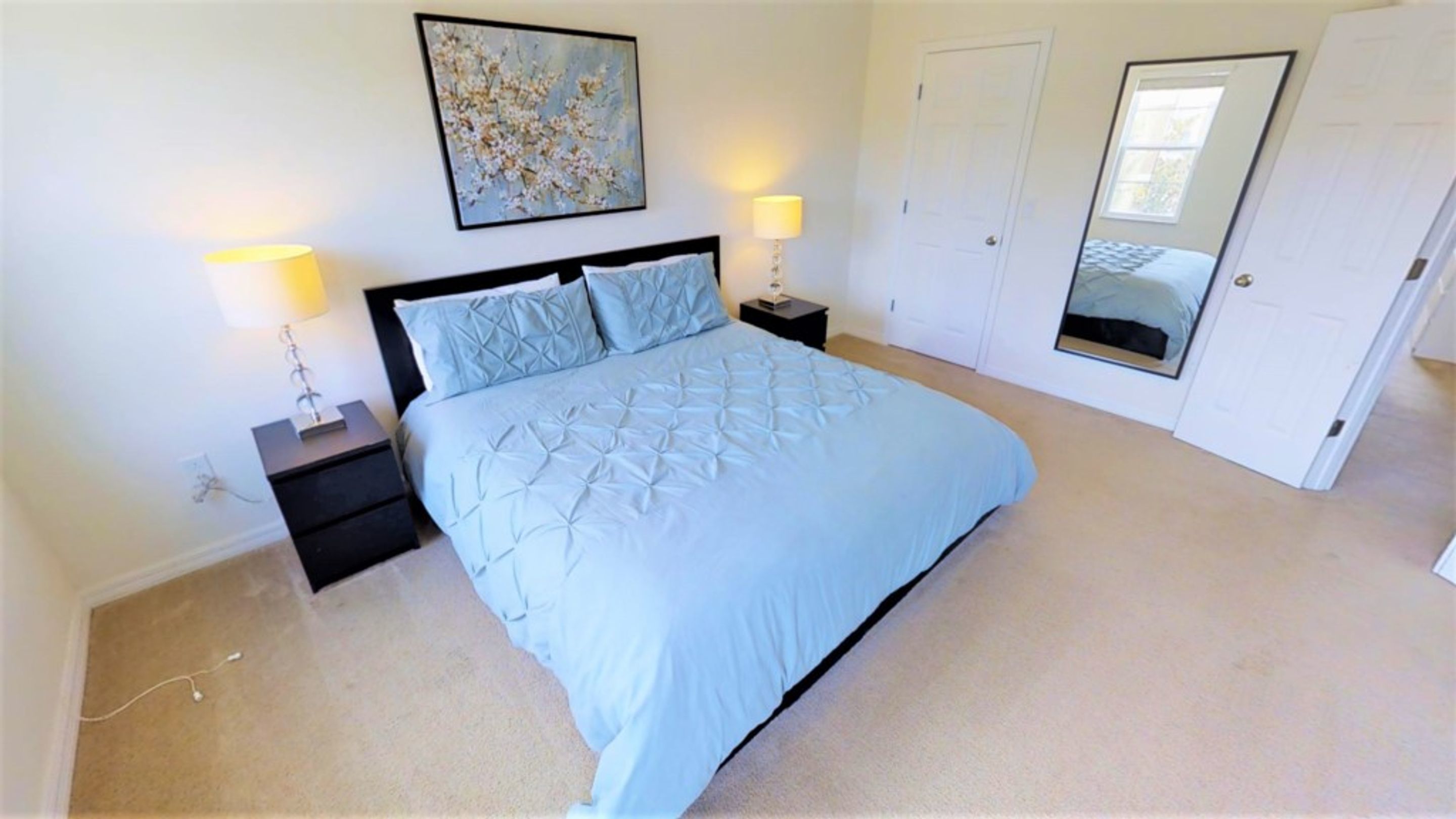 A bedroom with a bed, two bedside tables, a nightstand, a painting on the wall, and a door.