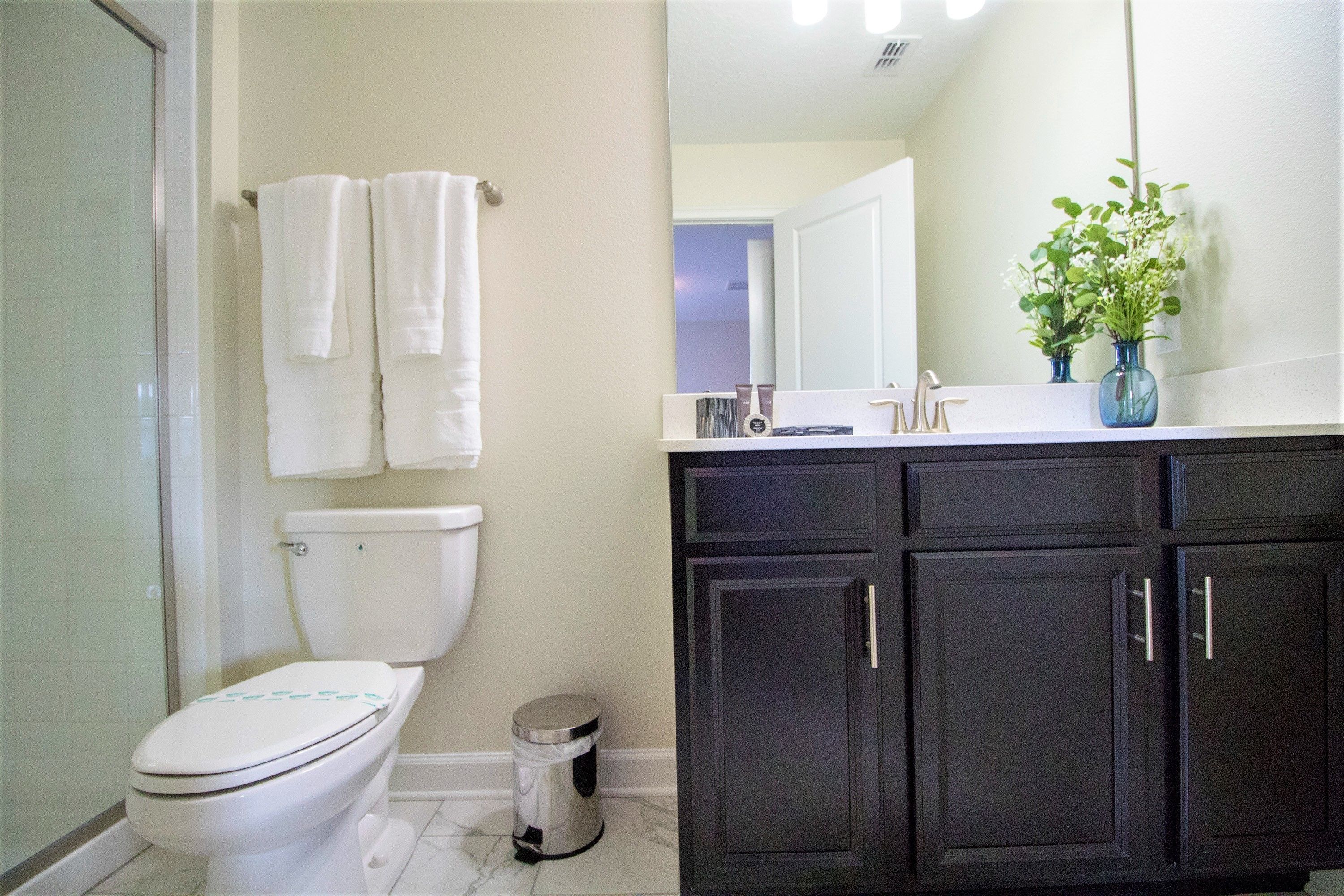 A bathroom with a toilet, a trash can, a shower, and dark cabinets.