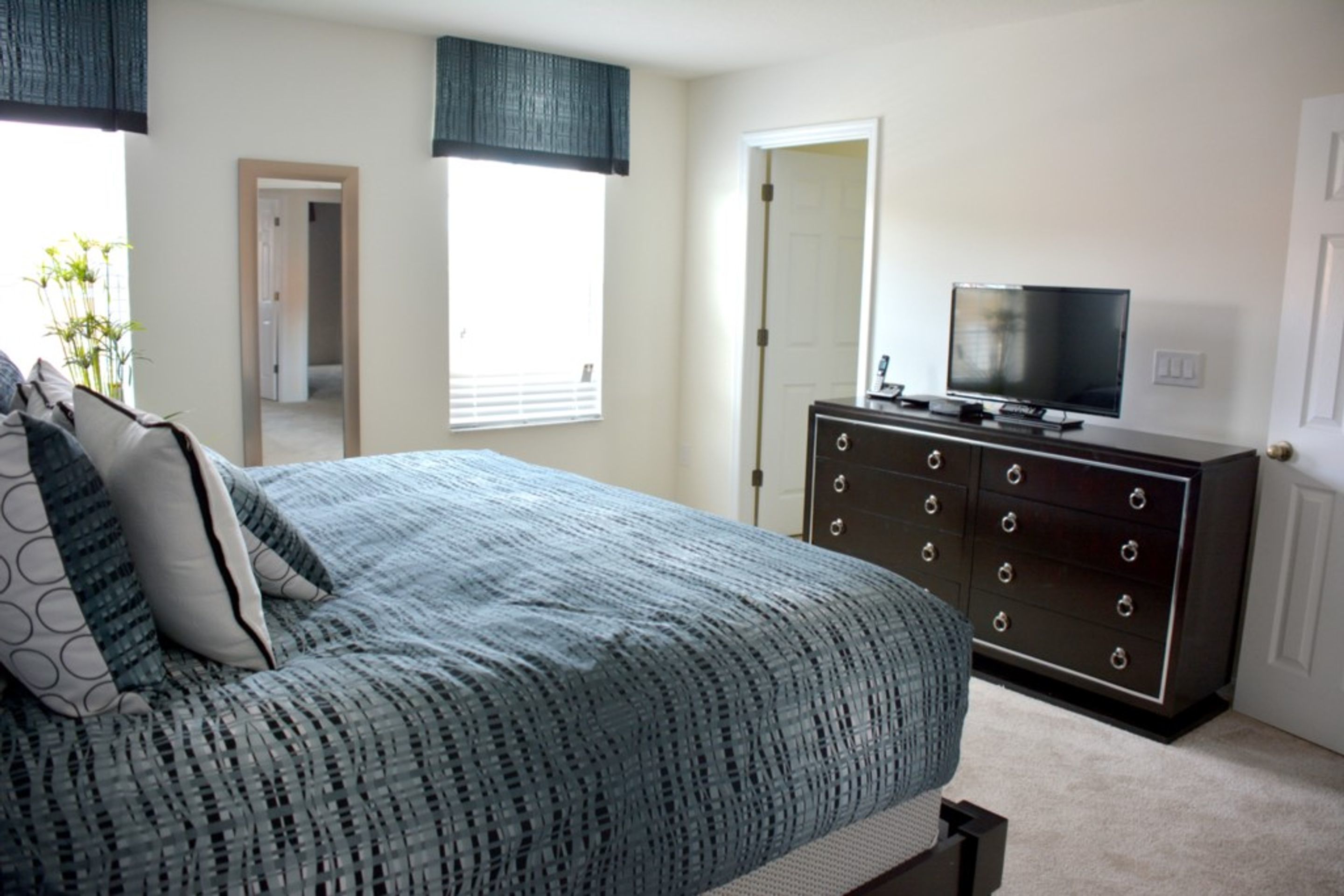 Une chambre à coucher comprenant un lit, une commode, un téléviseur et une fenêtre avec des stores.