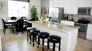 A modern kitchen with white cabinets, a stainless steel refrigerator, a microwave, and a coffee maker. There are four black bar stools and a dining table with chairs. - Bella Vida 851 (Kissimmee)