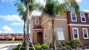 A two-story residential building with a beige exterior and red roof tiles. There are palm trees in front of the building, and a clear blue sky with some clouds in the background. - Bella Vida 851 (Kissimmee)