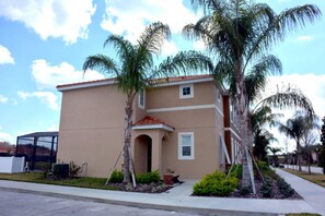 A two-story building with a beige facade, a red-tiled roof, and a small balcony. There are palm trees and shrubs in the front yard. - Bella Vida 851 (Kissimmee)